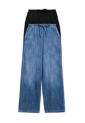 Kvinde iført hvid kortærmet crop top, vidde blå denim bukser med snørelukning i taljen, hvide sneakers og gyldt armbånd, stående mod en ensfarvet baggrund.