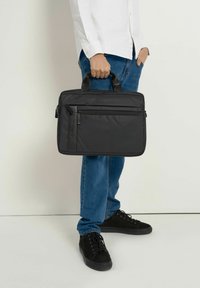 Czarna torba na laptopa z nylonu, z dwoma uchwytami, przednią kieszenią na zamek błyskawiczny i gładką fakturą. Trzymana przez osobę w białej koszuli i niebieskich dżinsach.