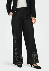 Črne široke hlače z elastnim pasom, okrašene z belim vezenjem cvetlic in palm po straneh. Tkanina z teksturo.