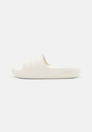 Enkelt cremefarvet slidesandal med teksturerede striber på remmen og flad sål på en ensfarvet hvid baggrund.