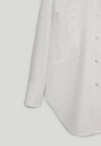 Biała koszula z długim rękawem, z guzikami z przodu, dużą kieszenią na piersi oraz wyprofilowanym, zaokrąglonym dołem na jasnym tle.