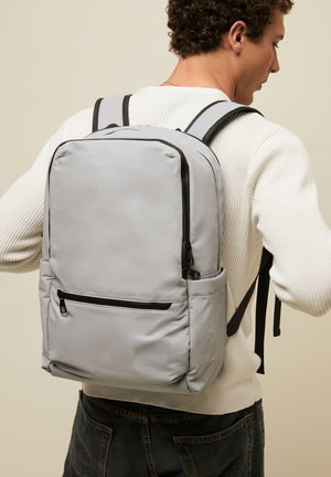 Muškarac nosi svijetlosivu ruksak s više džepova na munje preko bijelog džempera i tamnih traperica.