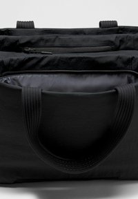 Melna iepirkumu soma ar platām dubultām rokturiem, kurā redzami atvērti nodalījumi un kabata ar rāvējslādzi iekšpusē.