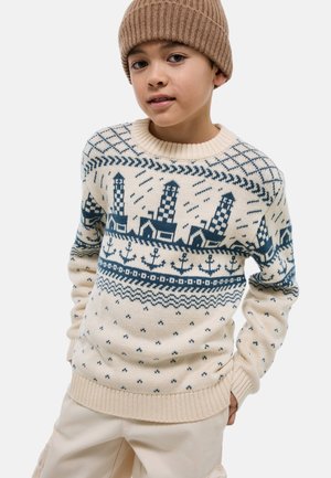 Garçon portant un pull en maille beige avec des motifs de phare et d'ancre, un pantalon beige et un bonnet côtelé marron, debout les mains dans les poches.