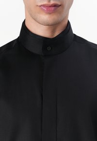 Μαύρο πουκάμισο με ψηλό γιακά, κλείσιμο με έναν μόνο κουμπί, μαλακή υφή και χωρίς ορατά σχέδια ή λεπτομέρειες. Κοντινό πλάνο στο άνω μέρος του κορμού.