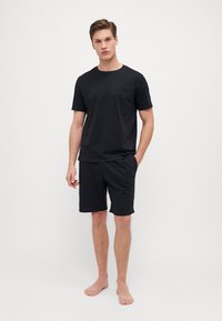 Schwarzes Baumwoll-T-Shirt mit einer Tasche, kombiniert mit schwarzen Shorts. Beide sind schlicht, bieten eine lockere Passform und weiche Textur. Das Model ist barfuß.