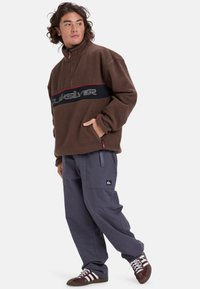 Sudadera de forro polar marrón con media cremallera, banda roja y negra que muestra "Quiksilver". Combinada con pantalones cargo gris oscuro y zapatillas marrones.