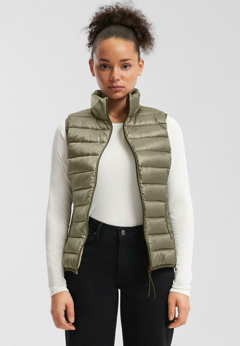 Eine Frau mit lockigem Haar trägt eine olivgrüne gesteppte Weste über einem weißen langärmligen gerippten Shirt und schwarzen hochgeschnittenen Hosen vor einem schlichten Hintergrund.