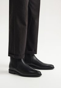 Czarne skórzane botki Chelsea z elastycznymi panelami bocznymi i gładkim wykończeniem, w połączeniu z ciemnymi, skróconymi spodniami. Prosty i elegancki design.