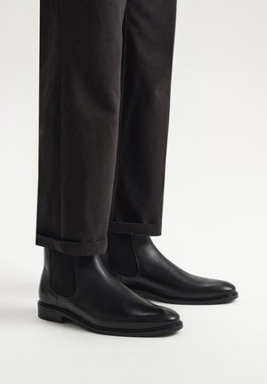 Zwarte leren Chelsea-boots met elastische zijpanelen en een gladde afwerking, gecombineerd met donkere, korte broeken. Eenvoudig en strak ontwerp.
