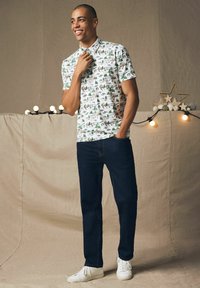 Kortærmet hvid skjorte med grøn og brun naturprint, parret med mørkeblå jeans og hvide sneakers. Enkelt design og rene linjer.