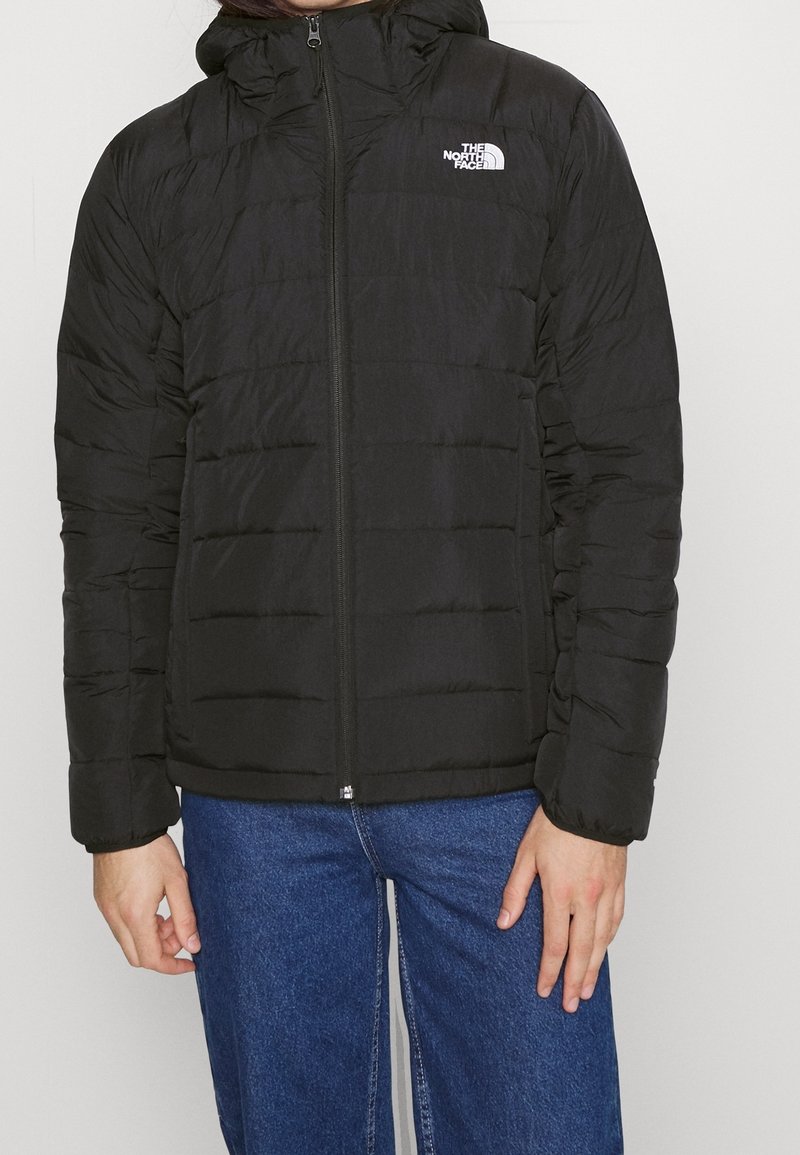 Personne portant une veste matelassée noire The North Face avec une fermeture éclair à l'avant et un jean bleu, debout sur un fond blanc.