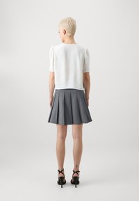 Weißes strukturiertes Kurzarmoberteil, kombiniert mit einem grauen, plissierten Rock. Das Model trägt schwarze High Heels, die ihre nackten Beine und ein Tattoo zur Geltung bringen.
