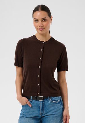 Donna con capelli scuri raccolti in una coda di cavallo indossa un cardigan marrone a maniche corte con bottoni di perla e jeans blu, in piedi contro uno sfondo semplice.
