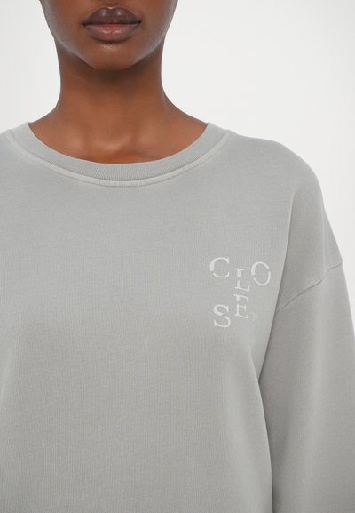 Felpa grigia con collo a giro e texture a costine. Presenta il testo ricamato bianco "CLO" e "SED" sul davanti. Vestibilità comoda.