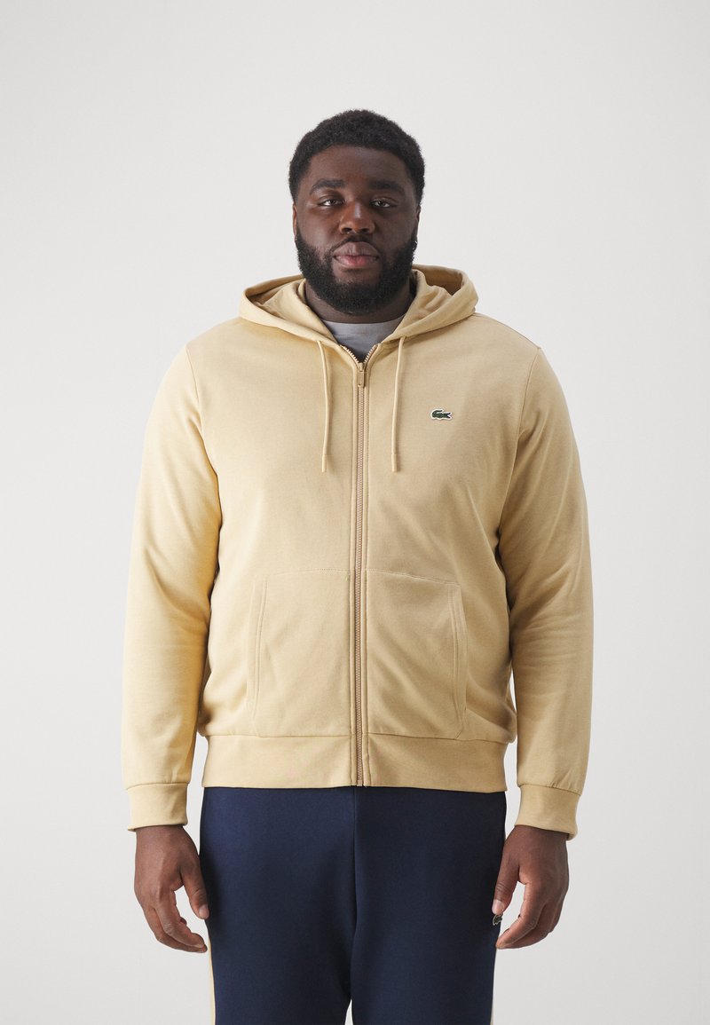 Beige ritsjack gemaakt van zachte stof, met een buidelzak, verstelbare capuchon en geborduurd groen logo op de linkerborst.