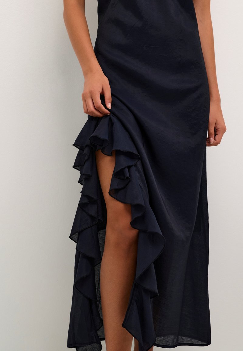 Navyblaues, satin Abendkleid mit einem rüschengesäumten Saum auf Oberschenkelhöhe und einem Schlitz, der das Bein enthüllt. Glatte Textur und leichtes Design.