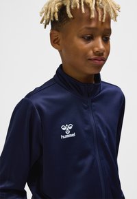 Navy sports jacket made of smooth fabric, featuring a high collar, zip closure, and a white logo on the chest.