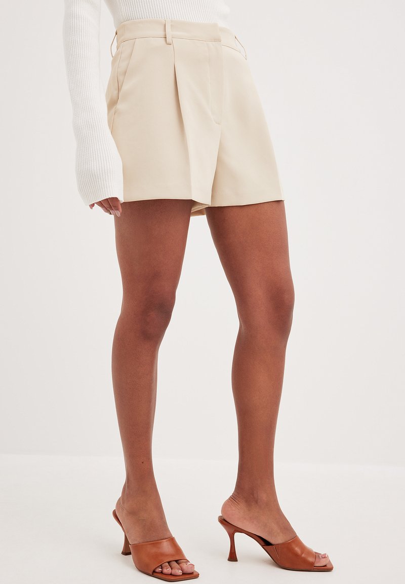 Beige lyhyet housut, joissa on laskoksia ja sivutaskut, valmistettu kevyestä materiaalista. Yhdistetty ruskeisiin avonaisiin korkokenkiin.