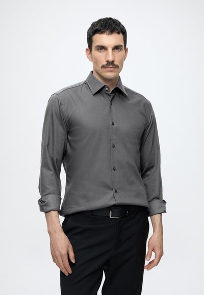 Homme aux cheveux foncés et moustache, portant une chemise grise boutonnée aux manches retroussées et un pantalon noir sur un fond clair uni.