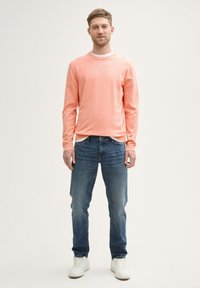 Helles Korallen-langärmliges Hemd mit Rundhalsausschnitt, kombiniert mit blauen Jeans und weißen Sneakers. Das Material wirkt weich und hat eine lässige Passform.