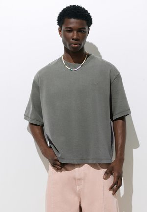 Jeune homme portant un t-shirt gris ample, un pantalon rose clair et un collier multicolore de perles, debout sur un fond blanc.