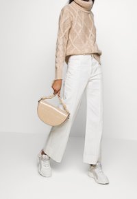 Beige gestrickter Rollkragenpullover mit Zopfmuster, weiße weit geschnittene Hose und eine strukturierte beigen halbkreisförmige Handtasche. Weiße Turnschuhe.