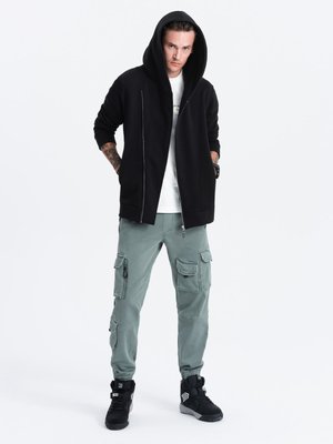 Zwarte rits hoodie met een grote capuchon, gecombineerd met lichtgroene cargobroeken met meerdere zakken en zwarte sportschoenen.