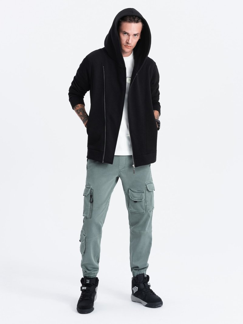 Zwarte rits hoodie met een grote capuchon, gecombineerd met lichtgroene cargobroeken met meerdere zakken en zwarte sportschoenen.