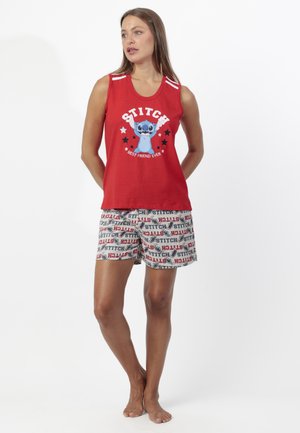 Joven mujer con camiseta sin mangas roja con el personaje Stitch y pantalones cortos beige con estampados de texto e imágenes de Stitch, de pie descalza.