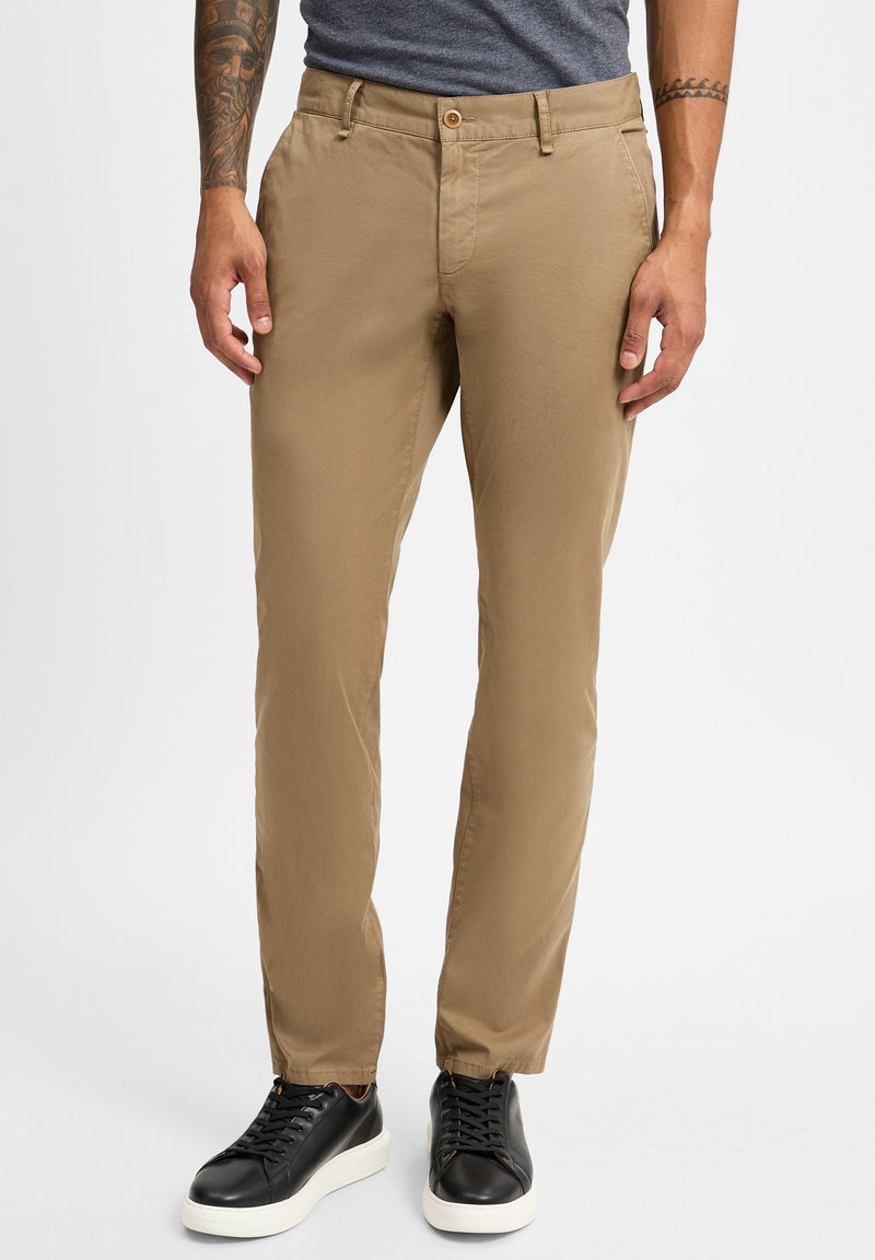 Beige Baumwollchinos mit glatter Textur, schmaler Passform und einem einzelnen Knopfverschluss. Kombiniert mit schwarzen Schnürsneakern mit weißer Sohle.