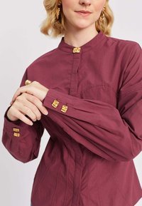 Donna che regola la manica di una blusa bordeaux con bottoni decorativi dorati e dettagli di cucitura minimi, mostrando il torso superiore e le mani.