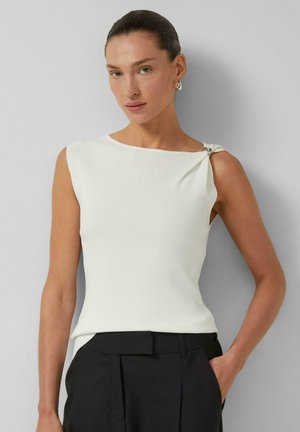 Femme aux cheveux plaqués en arrière, portant un haut blanc sans manches avec un nœud à l'épaule et un pantalon noir, debout contre un mur gris clair.