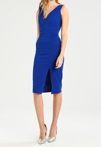 Robe ajustée bleue avec un profond décolleté en V, longueur au genou et une fente à l'avant. Fabriquée en tissu lisse, associée à des chaussures à talons hauts argentées.