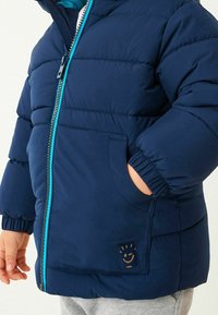 Veste matelassée bleu marine avec une fermeture éclair turquoise, présentant un design matelassé, des poignets élastiques et un patch logo smiley sur la poche avant.