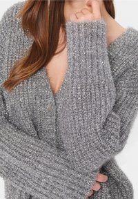 Šedý pletený cardigan s V-výstřihem, dlouhými žebrovanými rukávy a jedním knoflíkovým zapínáním. Materiál má třpytivý povrch a jemnou texturu.