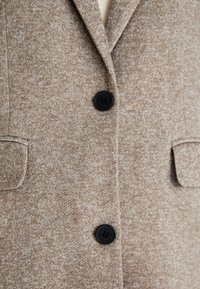 Manteau beige texturé avec deux boutons noirs et deux poches à rabat à l'avant, avec un col cranté.