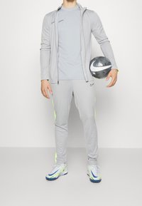 Conjunto deportivo gris con acentos en neón, que incluye una sudadera con cremallera, camiseta ceñida y pantalones. Incluye un balón de fútbol en la mano del sujeto.