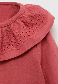 Lähikuva pehmeästä, ruosteenvärisestä kankaasta, jossa on pitsireunainen kaulus, jossa on kukka-ompelua ja läpivientimuotoiluja.