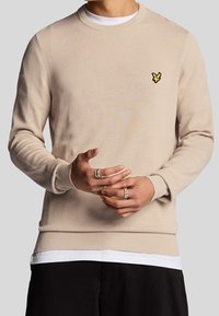 Beige knitted sweater featuring a crew neckline, ribbed cuffs, and hem, with a small yellow embroidered logo on the left chest.