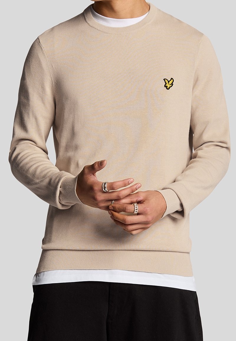 Beige knitted sweater featuring a crew neckline, ribbed cuffs, and hem, with a small yellow embroidered logo on the left chest.
