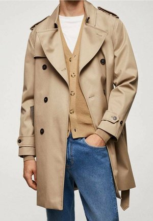 Beige dobbeltradet trenchcoat båret over en tan cardigan og hvid skjorte, kombineret med blå jeans, den ene hånd i lommen.