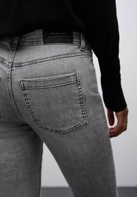 Los jeans de denim gris presentan un diseño ajustado, un bolsillo trasero con una solapa puntiaguda y una etiqueta negra en la cintura. La textura es suave y sutil.