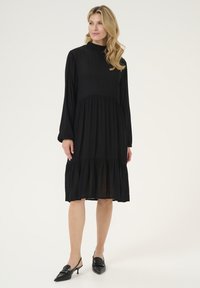Black long-sleeve dress with a high neck, tiered ruffle hem, and textured fabric. Paired with black slingback heels.