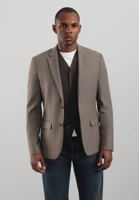 Jasnobrązowa marynarka o dopasowanym kroju, z dwoma przednimi kieszeniami i klapami z wycięciami, nałożona na ciemnobrązowy sweter rozpinany i białą koszulę.