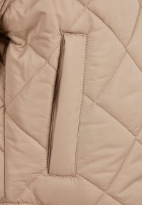 Beige gesteppter Jackenstoff mit einer flachen Tasche und genähtem Rand. Die Textur ist glatt mit einem subtilen Glanz und geometrischem Muster.