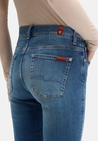 Niebieskie dżinsy z denimu o dopasowanym kroju, z wysokim stanem, dwoma tylnymi kieszeniami oraz czerwoną metką z cyfrą „7” na pasku w talii.