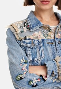 Chaqueta de mezclilla con bordado floral en los hombros y las mangas, textura desgastada, color azul claro y cierres de botón.
