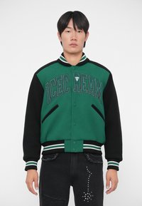 Giacca varsity verde e nera con la scritta "ICECREAM", bottoni a pressione, colletto e polsini a costine, e due tasche laterali.