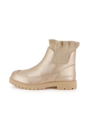 Bottine beige à la cheville avec bord côtelé en tricot, semelle plate et bout rond, présentant des panneaux latéraux élastiques et un marquage textuel discret sur la zone du talon.